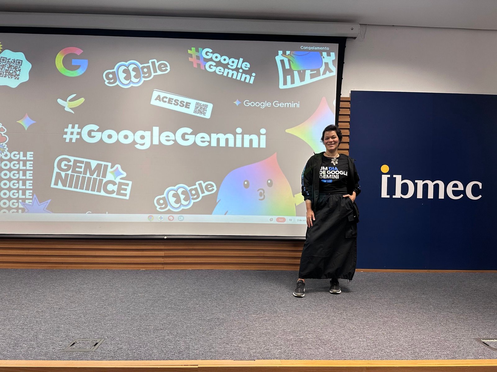 Roberta-Freitas_Empreender-na-educacao_3 Roberta Freitas em pé em um auditório, sorrindo, vestindo uma camiseta preta com os dizeres Um dia de Google Gemini sobre uma saia longa preta. Ao fundo, um telão coberto de adesivos e logos do Google Gemini, e à direita o logo do Ibmec.
