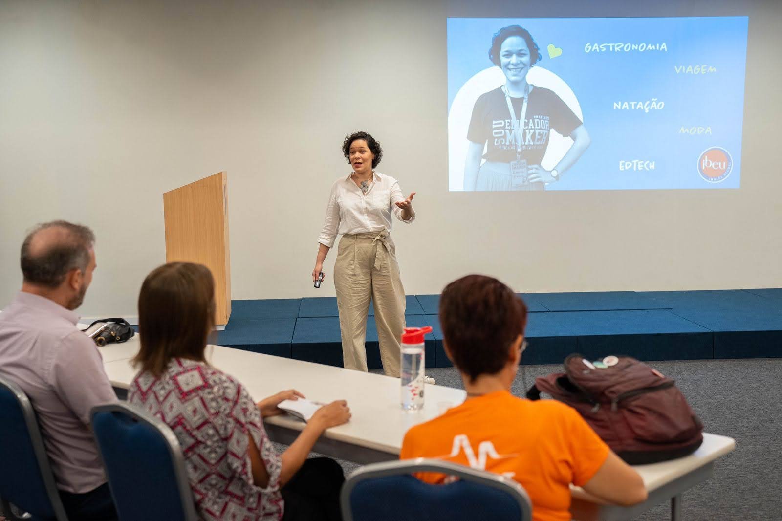 Roberta-Freitas_Empreender-na-educacao_4 Roberta Freitas em pé, de blusa branca e calça bege, gesticulando durante uma palestra em sala de aula. Ao fundo, um telão mostra uma foto dela em preto e branco vestindo uma camiseta com os dizeres Sou educador maker, cercada pelas palavras gastronomia, viagem, natação, moda e edtech, com o logo do IBEU.