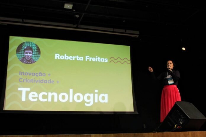 Roberta Freitas em pé em um auditório escuro, segurando um microfone e gesticulando, vestindo blusa preta e saia vermelha. Ao fundo, um telão verde projeta um slide com o nome dela e os dizeres Inovação mais Criatividade mais Tecnologia.